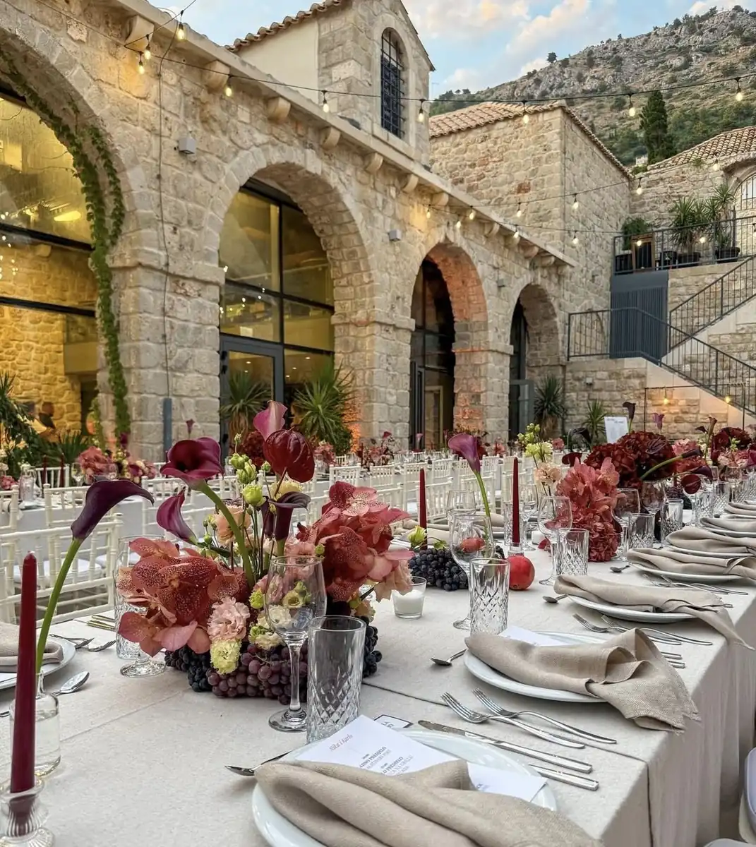 Longue table de dîner dressée sous des arcades en pierre, avec fleurs bordeaux et bougies
